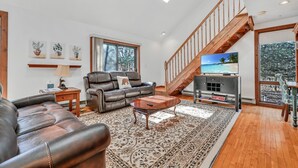 A spacious living room with a leather sofa, a coffee table, and a TV on a wooden stand. A staircase leads to an upper level, and there are large windows providing natural light. - Jim Thorpe, Bear Creek Lake, Cozy 3Br family friendly home- EV charging outlet (Jim Thorpe)