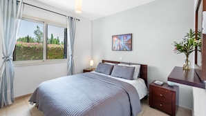 A bedroom with a large bed, a nightstand, a bedside table, a window with curtains, and a painting on the wall.