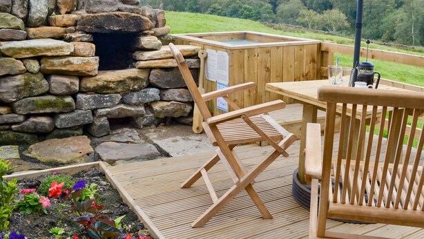 A wooden deck with a stone fireplace, a wooden table, and a chair. - The Old Back Kitchen at Bonfield Ghyll Farm (Helmsley)
