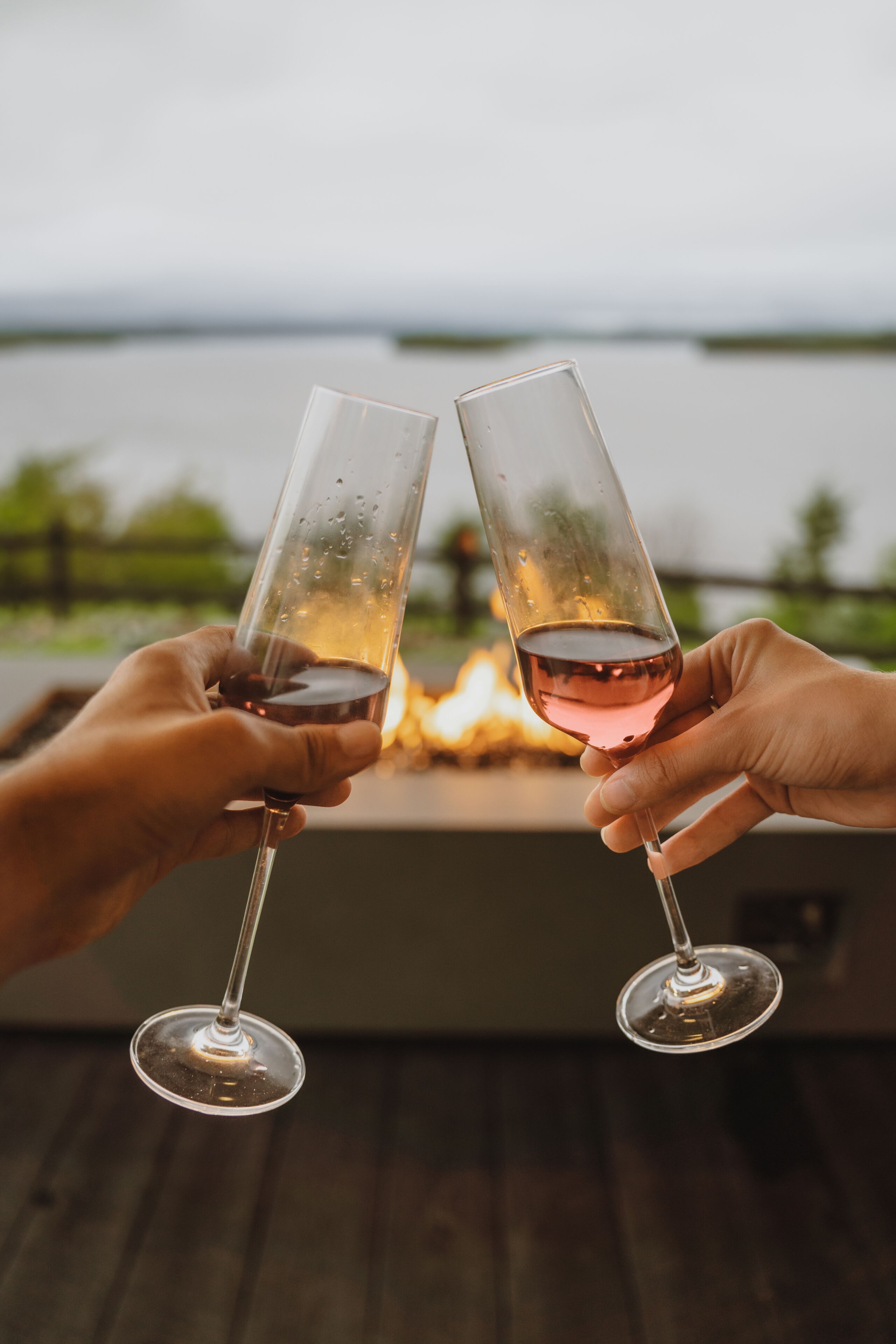 Duas mãos segurando copos de vinho com uma lareira ao fundo.