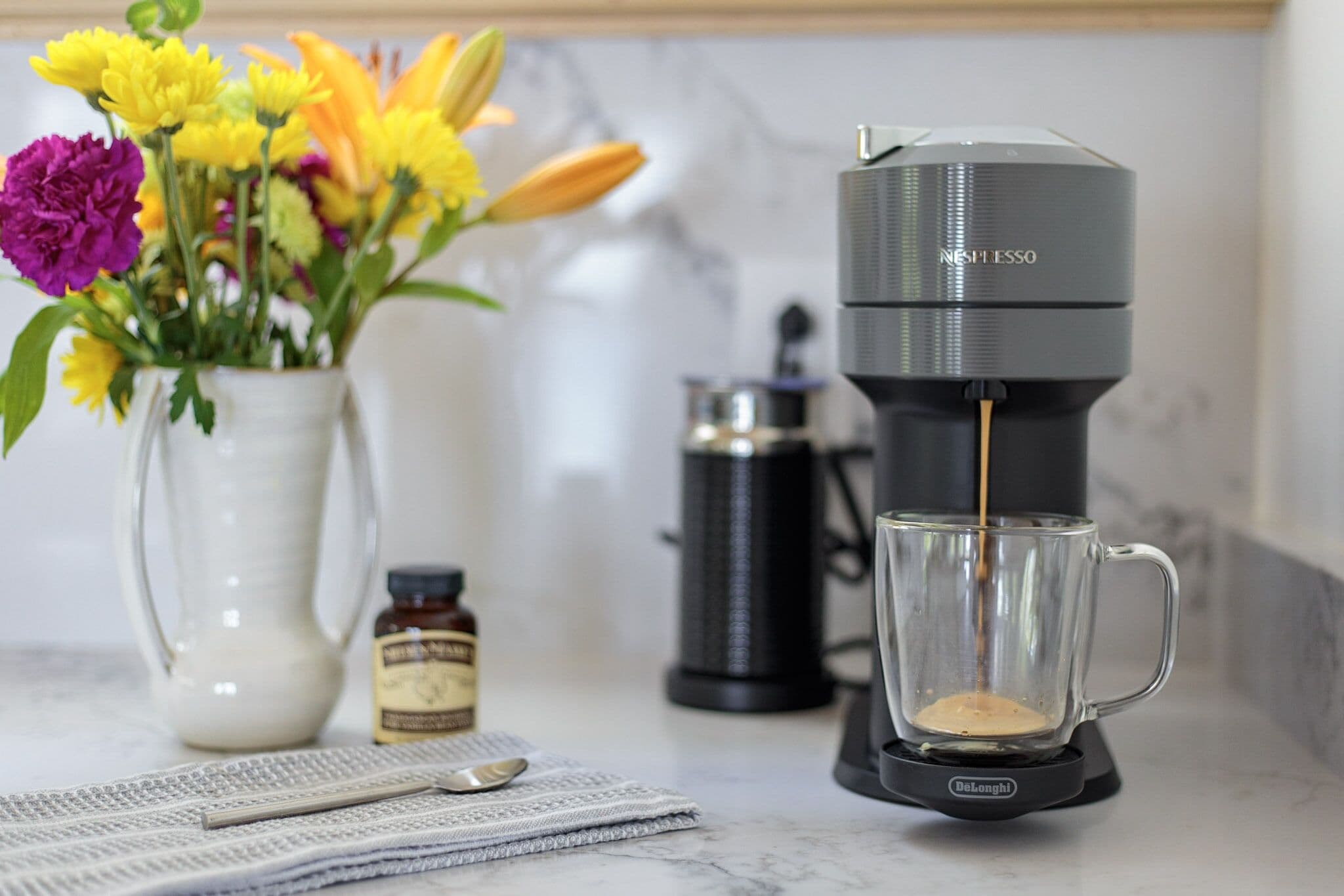 A coffee maker with a clear glass mug on a marble countertop, a white vase with flowers, and a small bottle of syrup.
