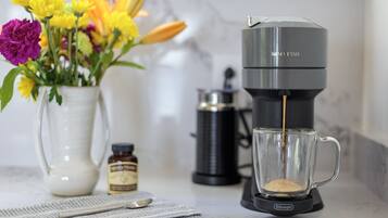 A coffee maker with a clear glass mug on a marble countertop, a white vase with flowers, and a small bottle of syrup.