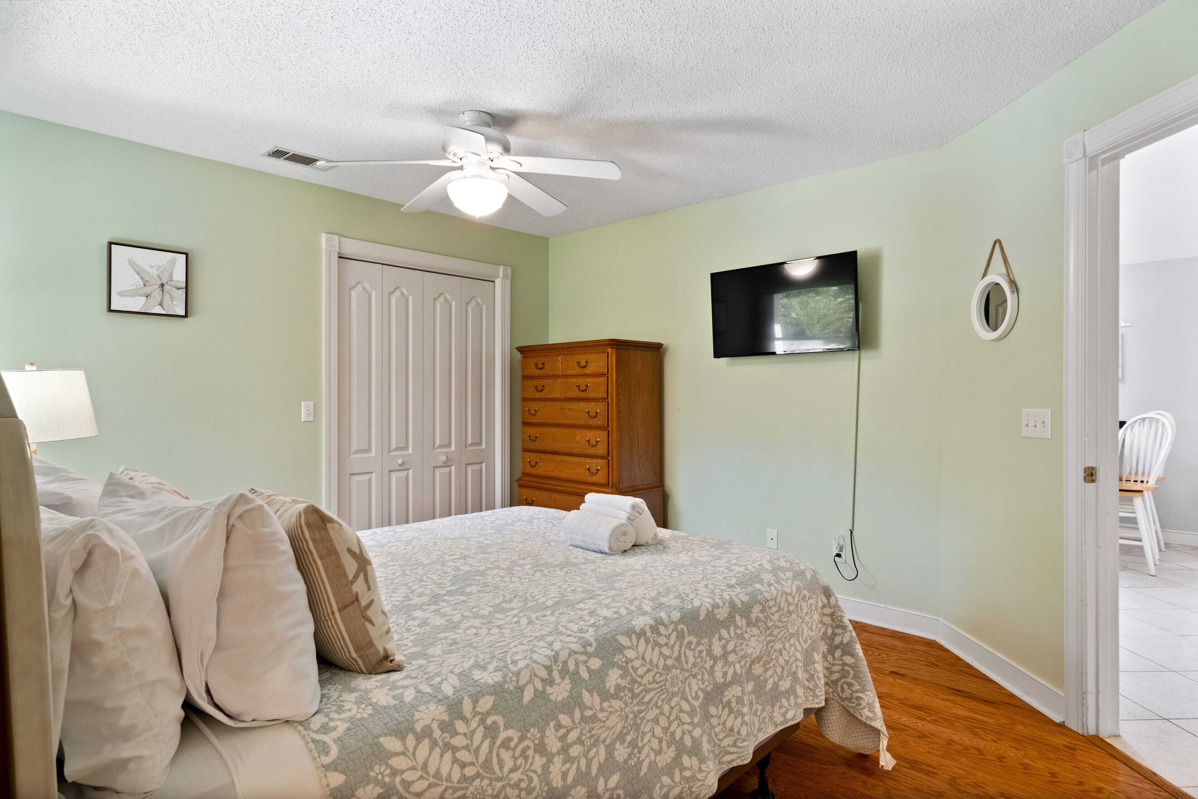 Un dormitorio con una cama, un ventilador de techo, un aparador, un televisor y una puerta que da a otra habitación.