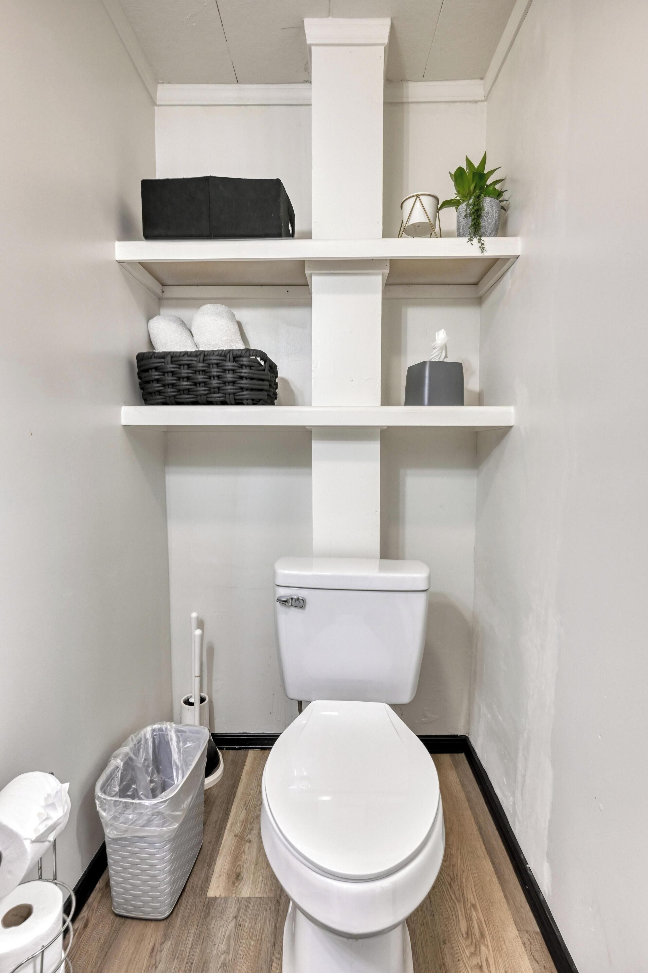 Une petite salle de bain avec des toilettes blanches, une poubelle, un panier et une étagère ornée d’une plante et d’une petite statue.