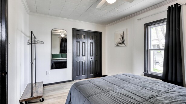 A bedroom with a bed, a ceiling fan, a door, a window with curtains, and a coat rack. - The Bluebird: Modern 1 Bed, Large Peaceful Yard (Joplin)