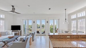Uma sala de estar espaçosa com uma televisão grande, assentos confortáveis, uma área de cozinha com bancadas de granito e grandes janelas com vista para o exterior.