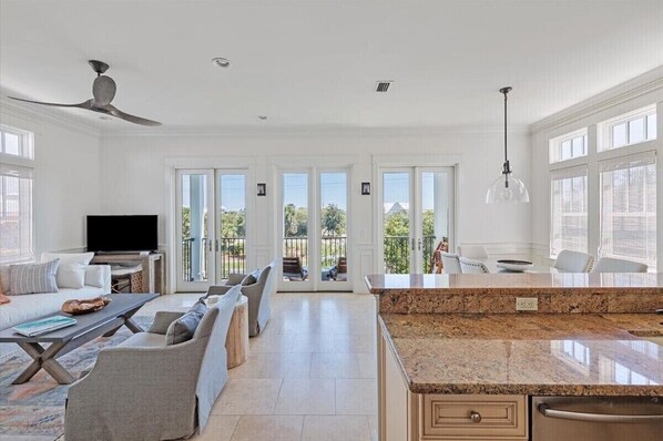 A spacious living room with a large TV, comfortable seating, a kitchen area with granite countertops, and large windows offering a view of the outdoors.