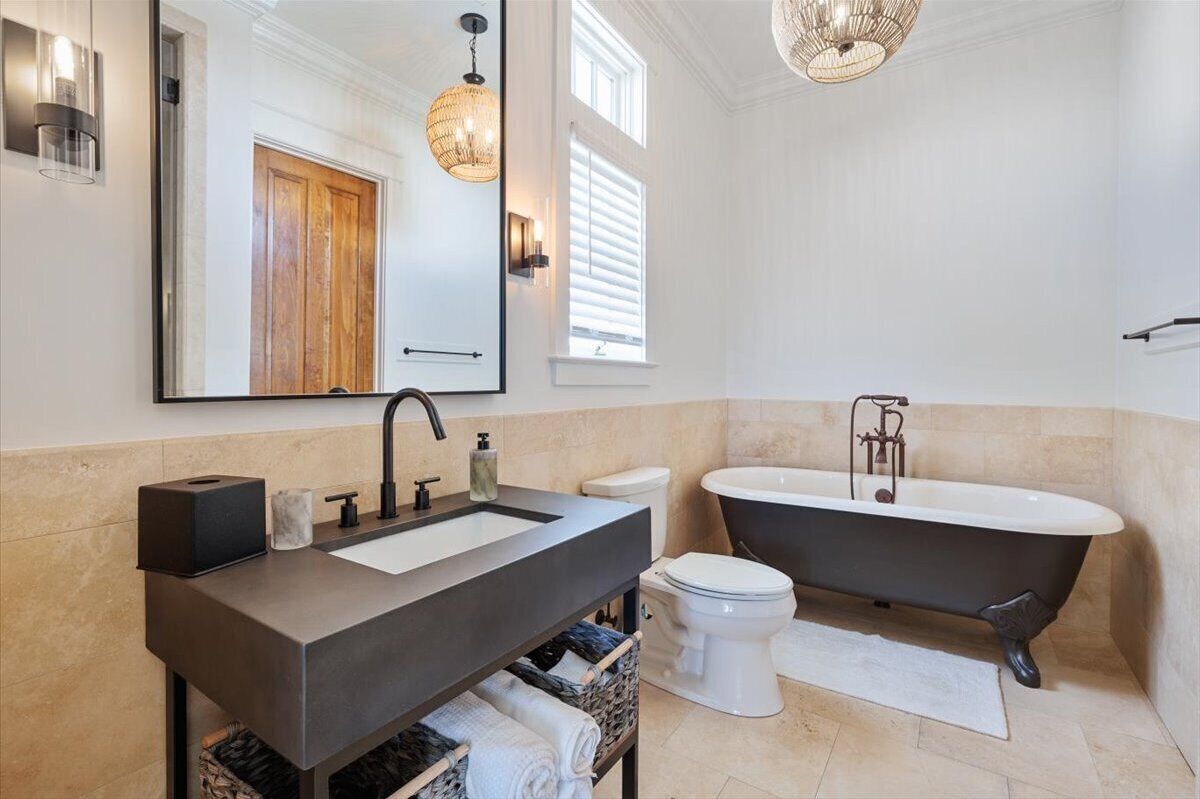 A modern bathroom with a large mirror, a dark sink, a white toilet, and a freestanding bathtub.