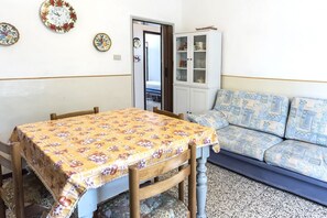 Una habitación con una mesa de madera cubierta por un mantel floral, un sofá azul con estampado, un armario blanco con puertas de cristal y una puerta que da a otra habitación.