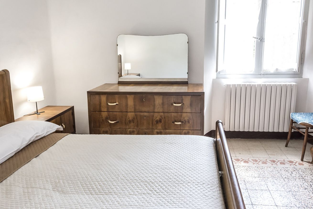 A bedroom with a bed, wooden dresser, mirror, bedside lamp, radiator, and window.