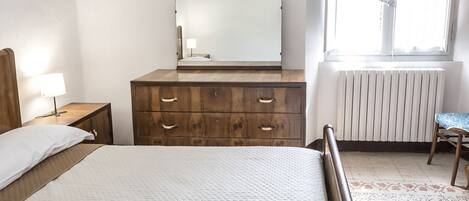 A bedroom with a bed, wooden dresser, mirror, bedside lamp, radiator, and window.