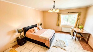 Un dormitorio con cama, mesitas de noche, un escritorio, una silla, un ventanal con cortinas y una araña.