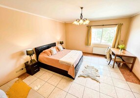 Un dormitorio con cama, mesitas de noche, un escritorio, una silla, un ventanal con cortinas y una araña.