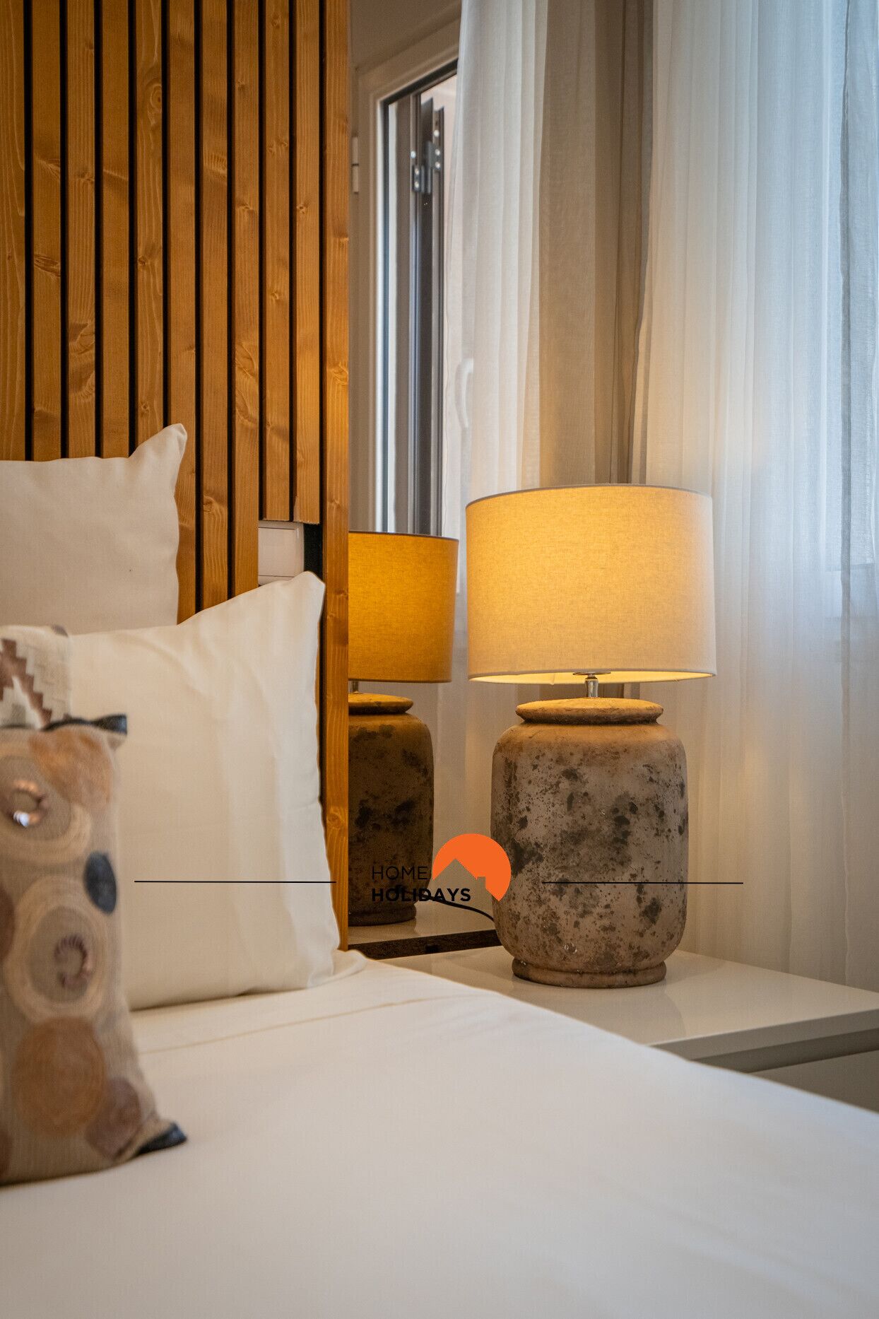 Una habitación de hotel con cabecera de madera, una cama con ropa de cama blanca, una mesita de noche con lámpara y un cojín decorativo.