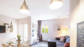 A modern living room with a sofa, a glass dining table, and a TV. There is a kitchen area with a fridge and a TV. - #094 Maresia Flats by Home Holidays (Albufeira)
