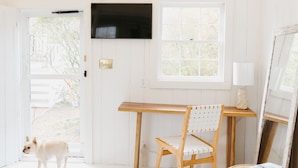 A bright, white room with a wooden desk, a chair, a small round table with books, a fluffy white rug, and a dog standing by a door. - Beach chic guest cottage, in town, just renovated (Nantucket)