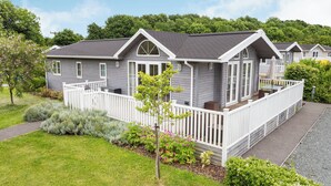 Une maison mobile d’un seul étage, de couleur grise, avec une véranda et une terrasse blanches, entourée d’une végétation luxuriante et sous un ciel dégagé.