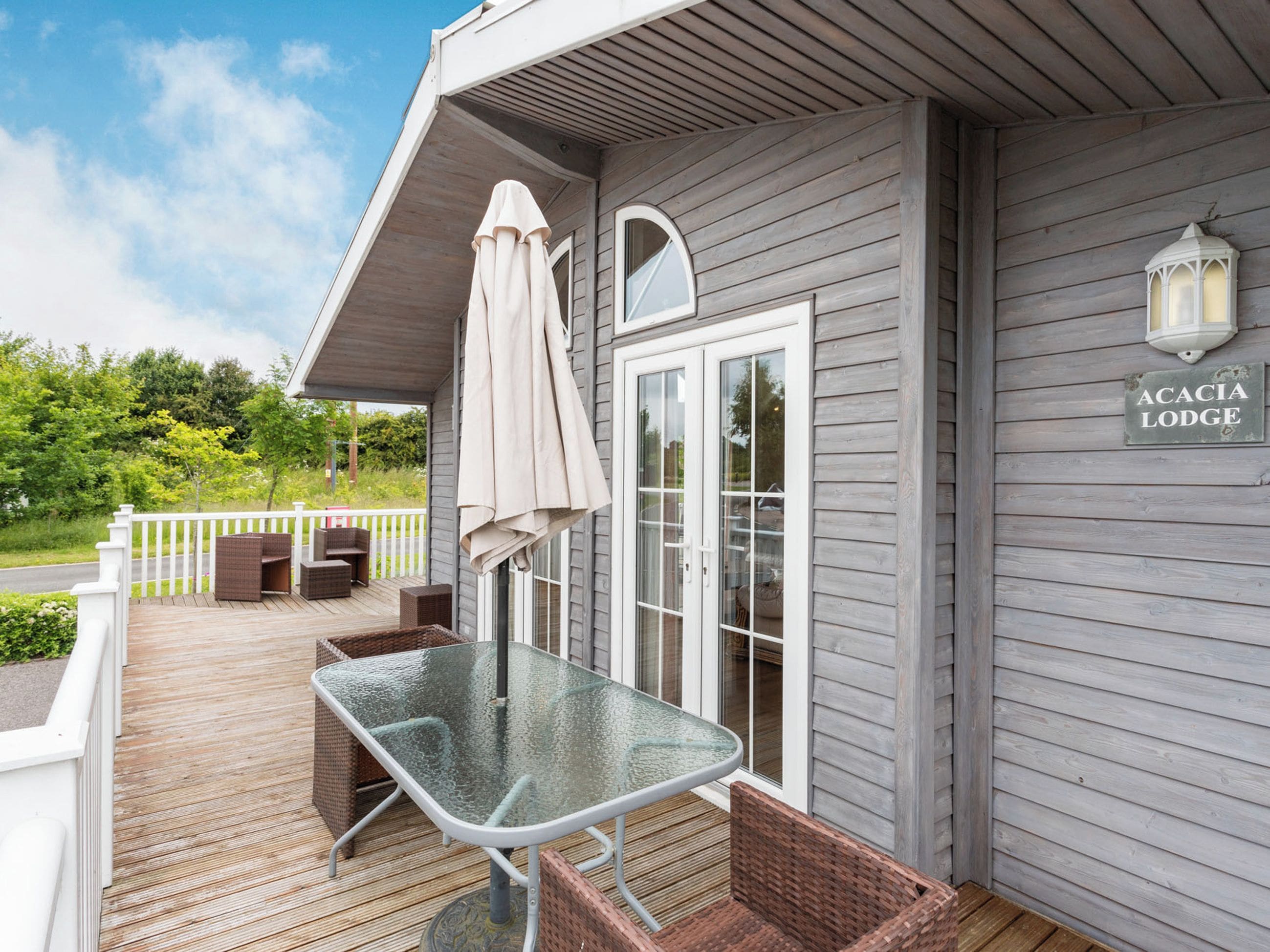 A wooden deck with a glass table and wicker chairs, a covered umbrella, and a sign reading "ACACIA LODGE".