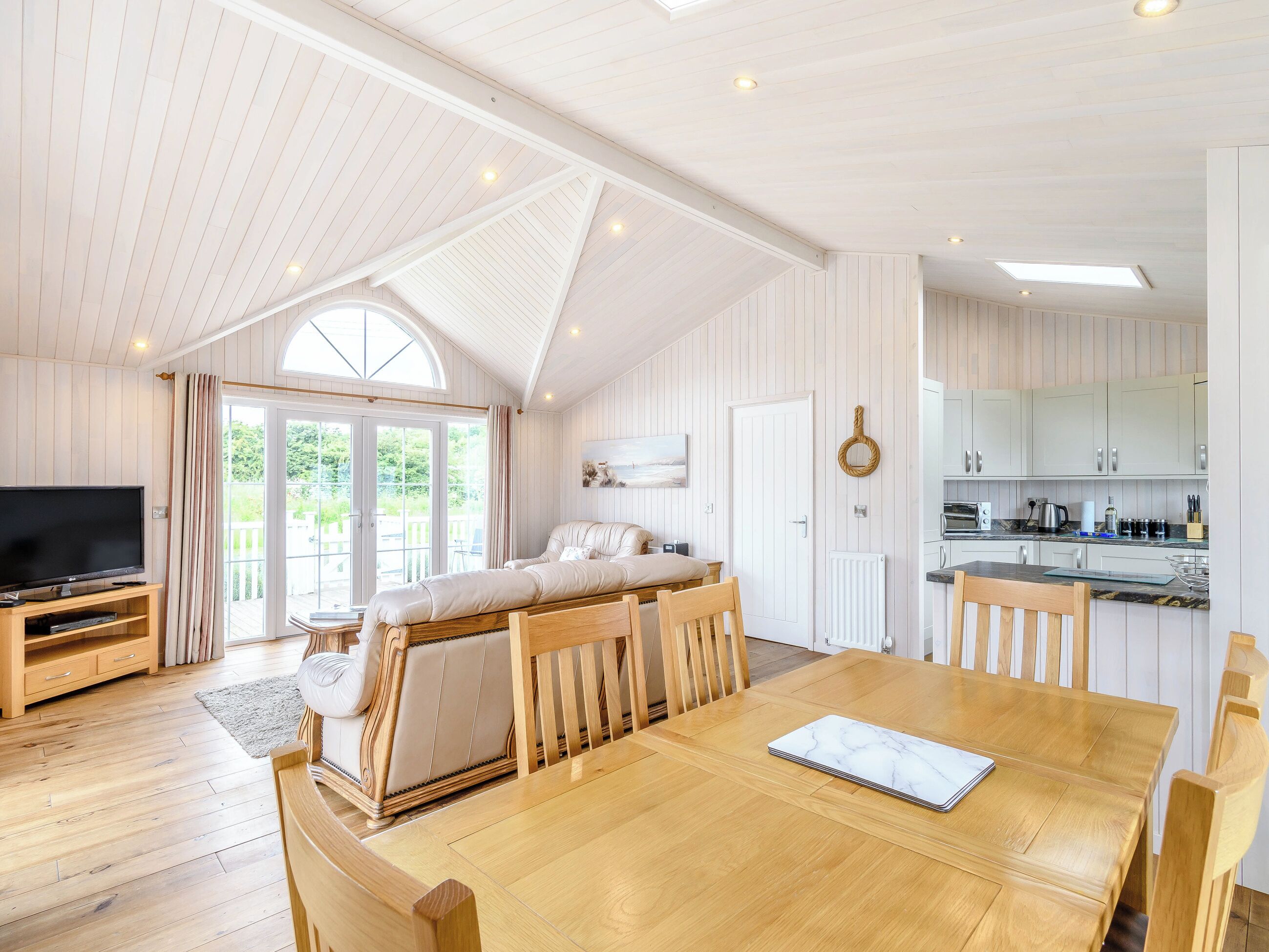 A spacious living area with a wooden dining table, a sofa, and a television. There is a kitchen area with white cabinets and a large window.