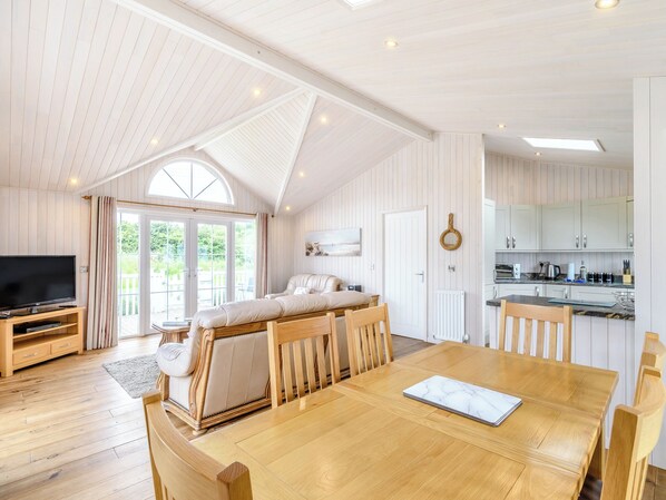 A spacious living area with a wooden dining table, a sofa, and a television. There is a kitchen area with white cabinets and a large window.