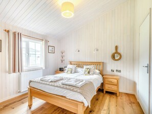 A bedroom with a wooden bed, bedside tables, a window with curtains, a wall-mounted light fixture, and a door with a round mirror.