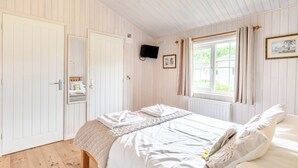Een slaapkamer met een bed, houten vloer, witte muren, een raam met gordijnen en een wandtelevisie.