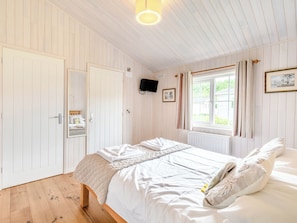 Un dormitorio con cama, suelo de madera, paredes blancas, una ventana con cortinas y un televisor de pared.
