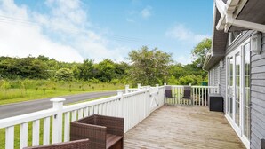 Une terrasse en bois, meublée de meubles en osier, avec une balustrade blanche et une vue sur une route et la verdure environnante.