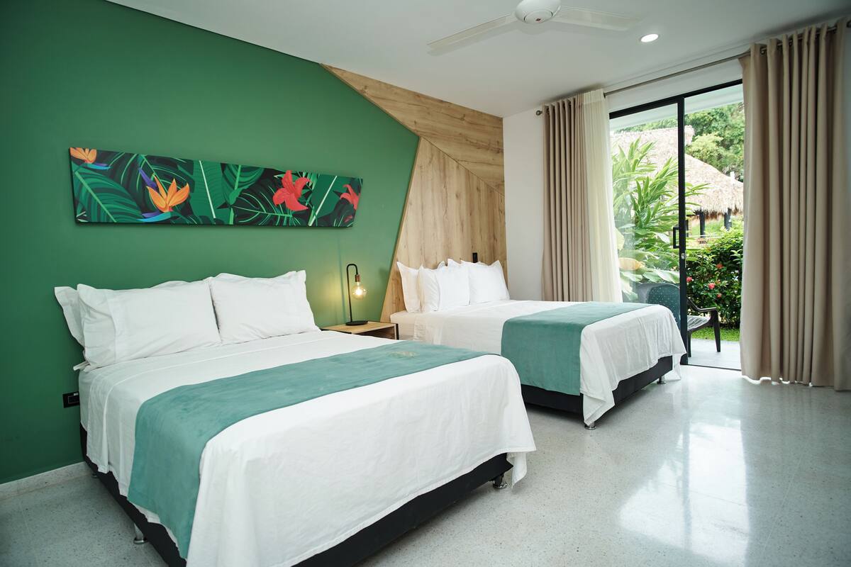 A hotel room with two beds, a green wall, a ceiling fan, and a view of a garden through a sliding door.