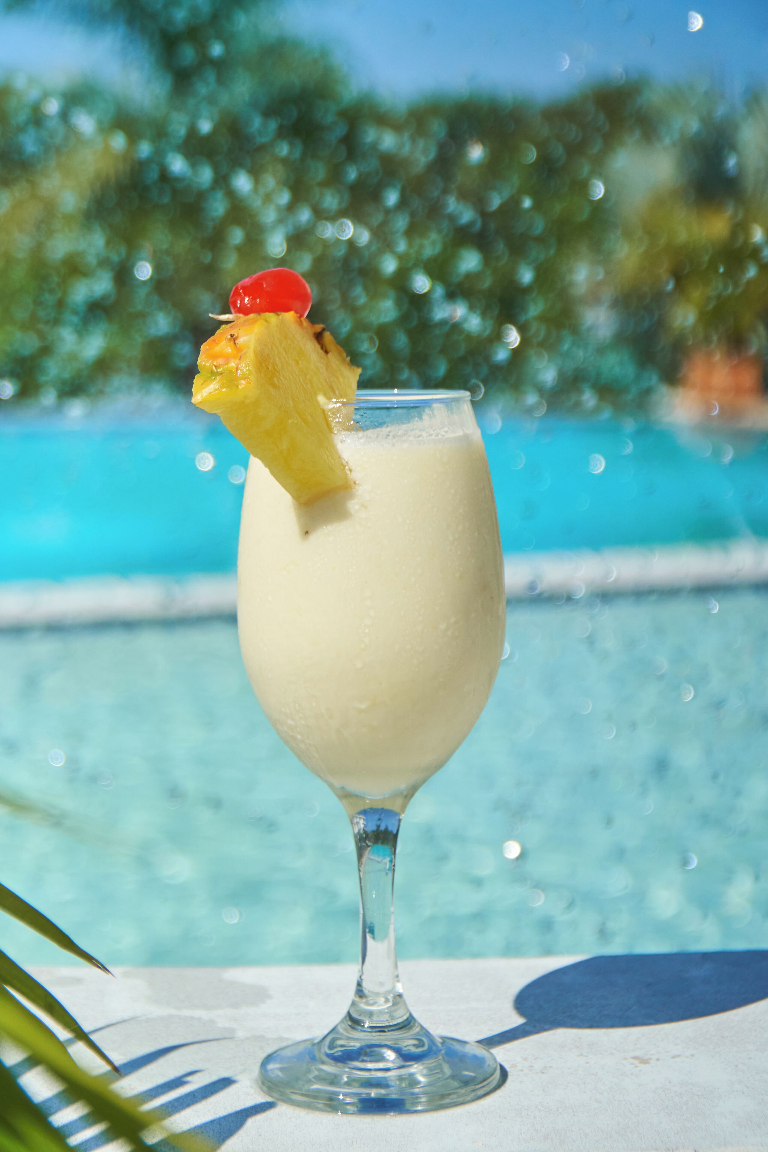 En cocktail med ananas og kirsebærpynt, serveret i et klart glas, ved en pool med uskarpt baggrund.