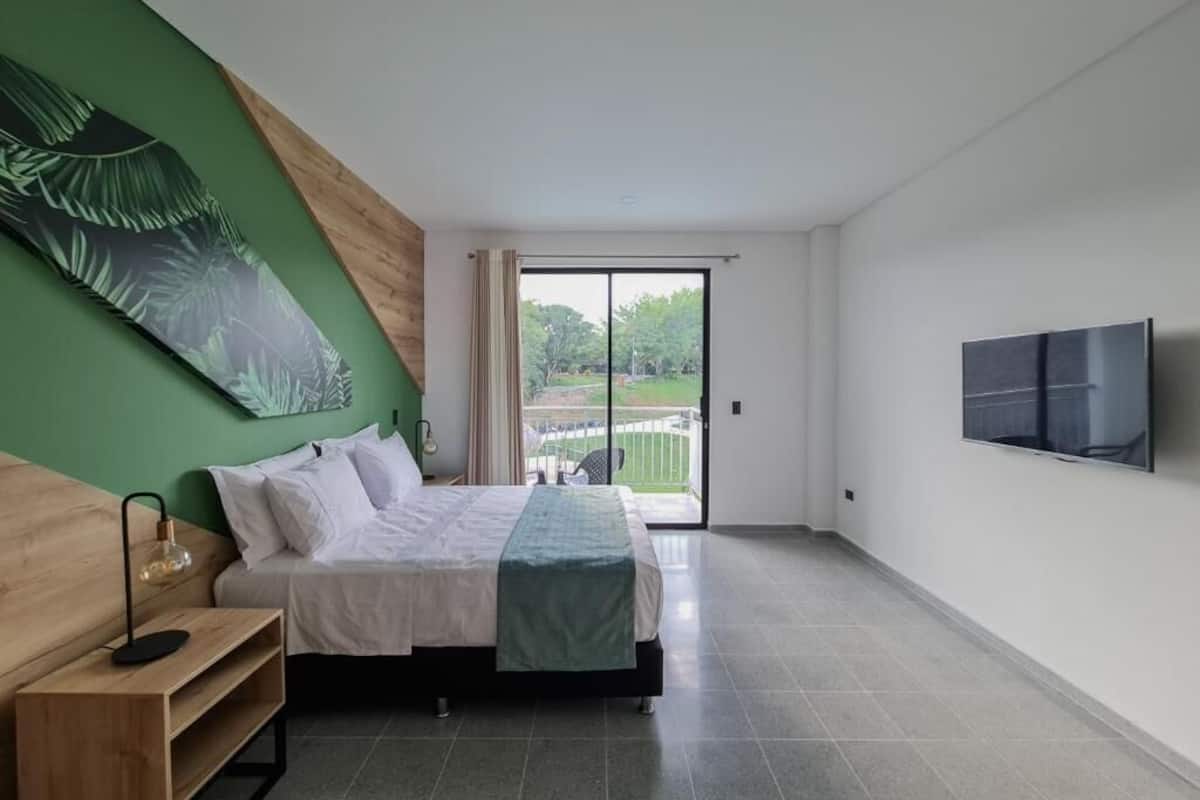 A bedroom with a bed, a nightstand, a lamp, a green wall art, a sliding glass door to a balcony, and a TV mounted on the wall.