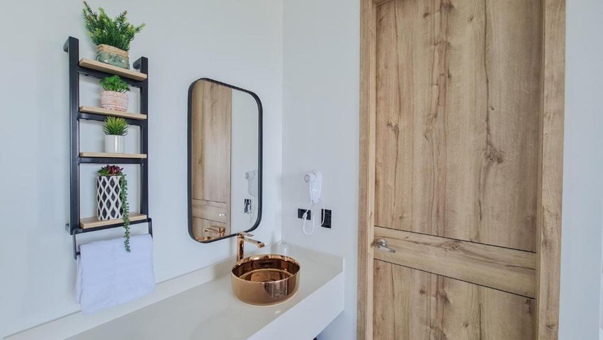 A bathroom with a white sink, a mirror, a towel rack with plants, a wooden door, and a hairdryer.