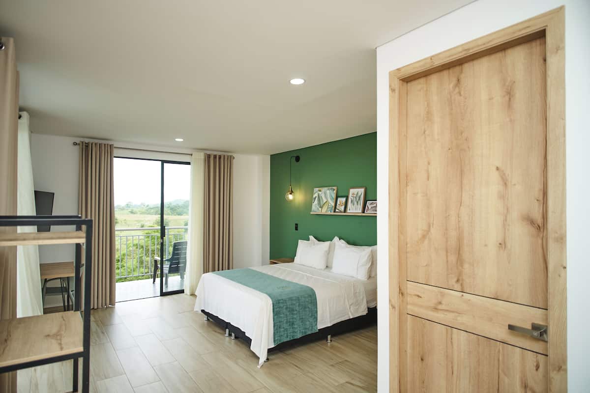 A bedroom with a bed, a desk, a chair, a balcony with a view, and a wooden door.