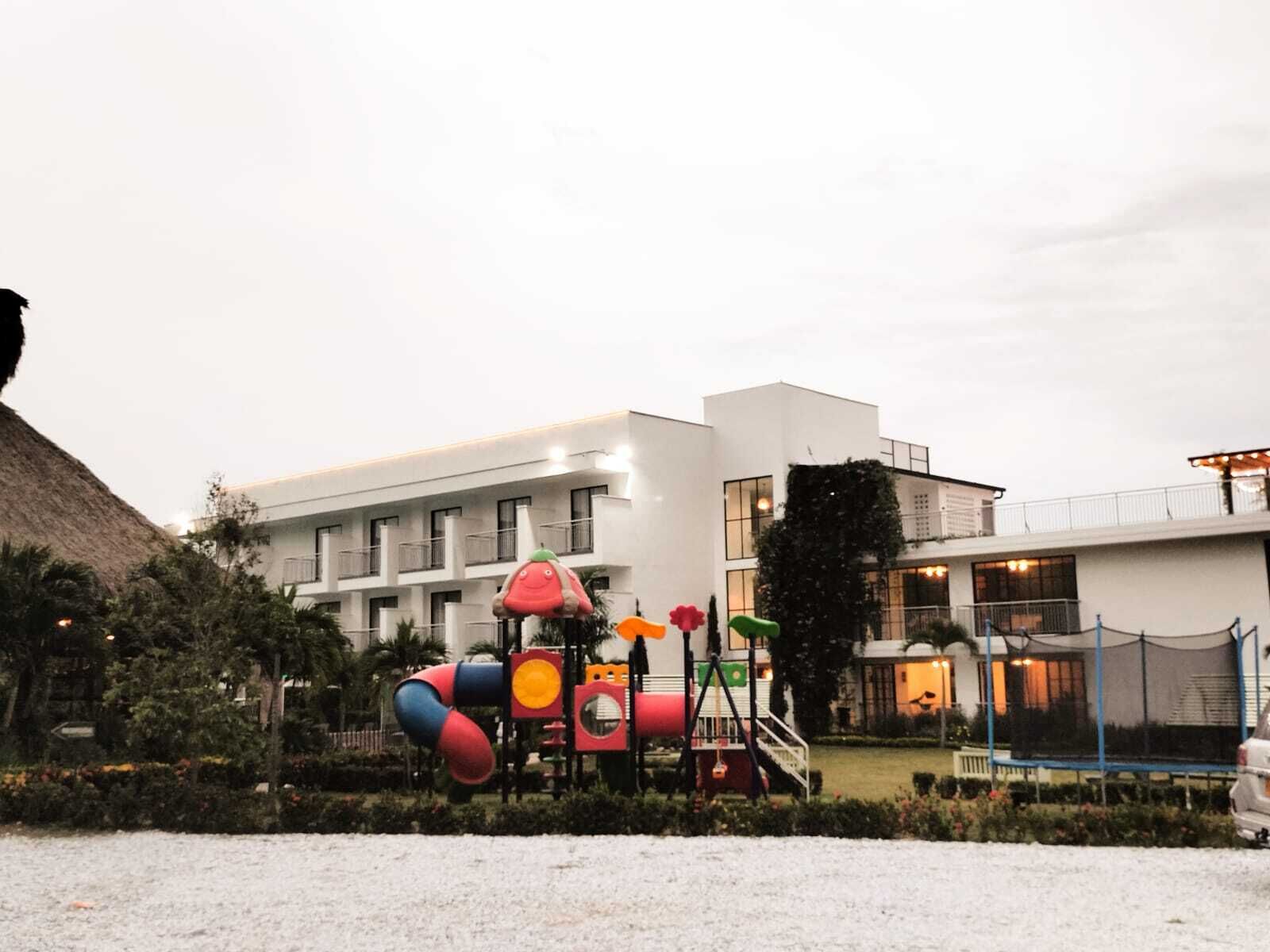 Ein Spielplatz mit bunten Geräten, ein weißes Gebäude mit vielen Fenstern und ein überdachter Bereich mit Beleuchtung.