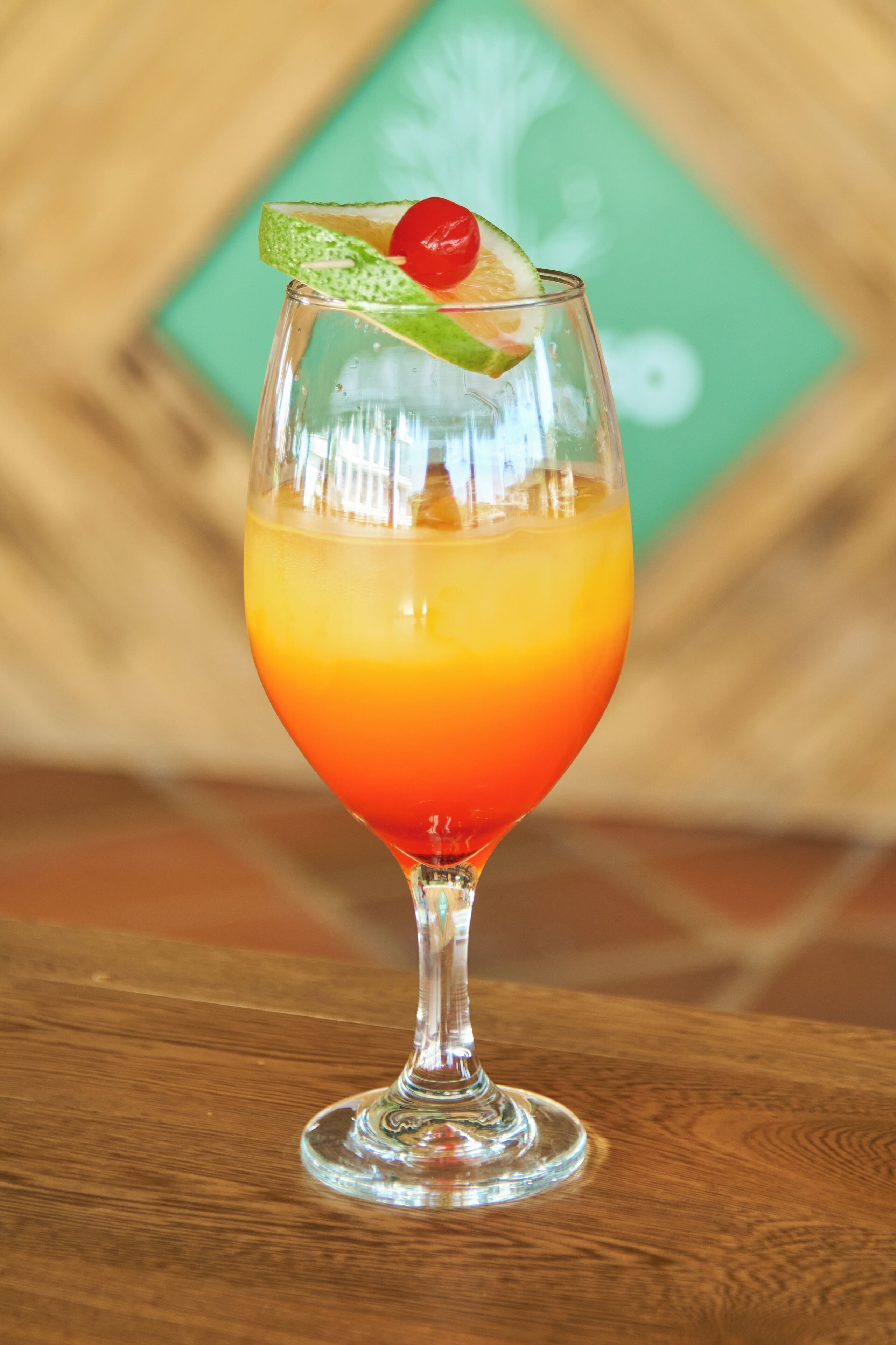 A cocktail glass with a gradient of orange and red, garnished with a lime slice and a cherry, on a wooden table.