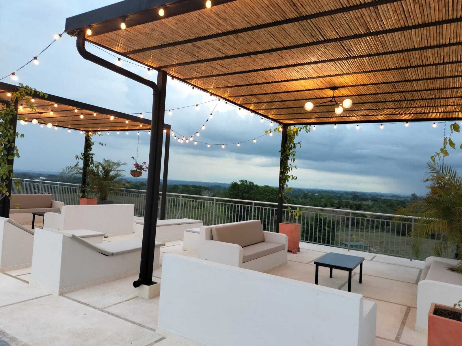 Un patio exterior con sofás blancos, una pérgola con luces colgantes y vistas al paisaje.