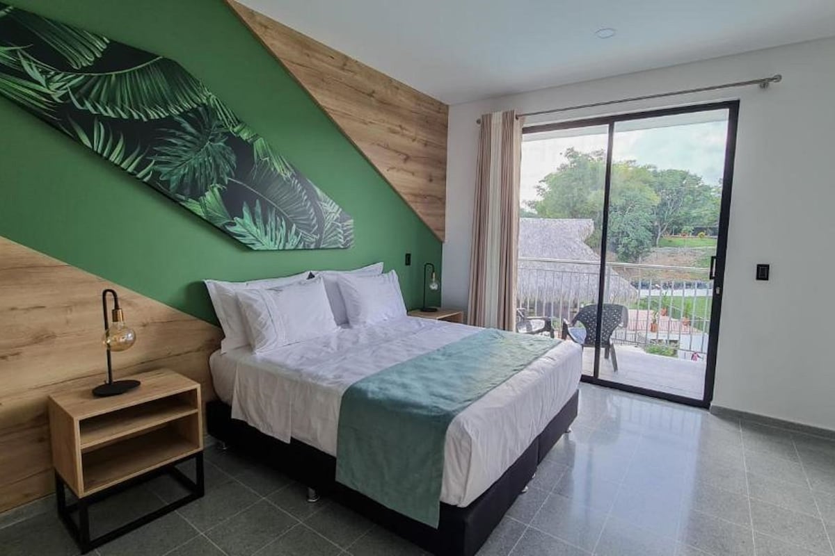 A bedroom with a bed, a nightstand, a green wall art, a window with curtains, and a view of the outdoors.