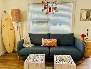 A cozy living room with a dark green sofa, two wooden side tables, a surfboard, a lamp, and a decorative hanging.