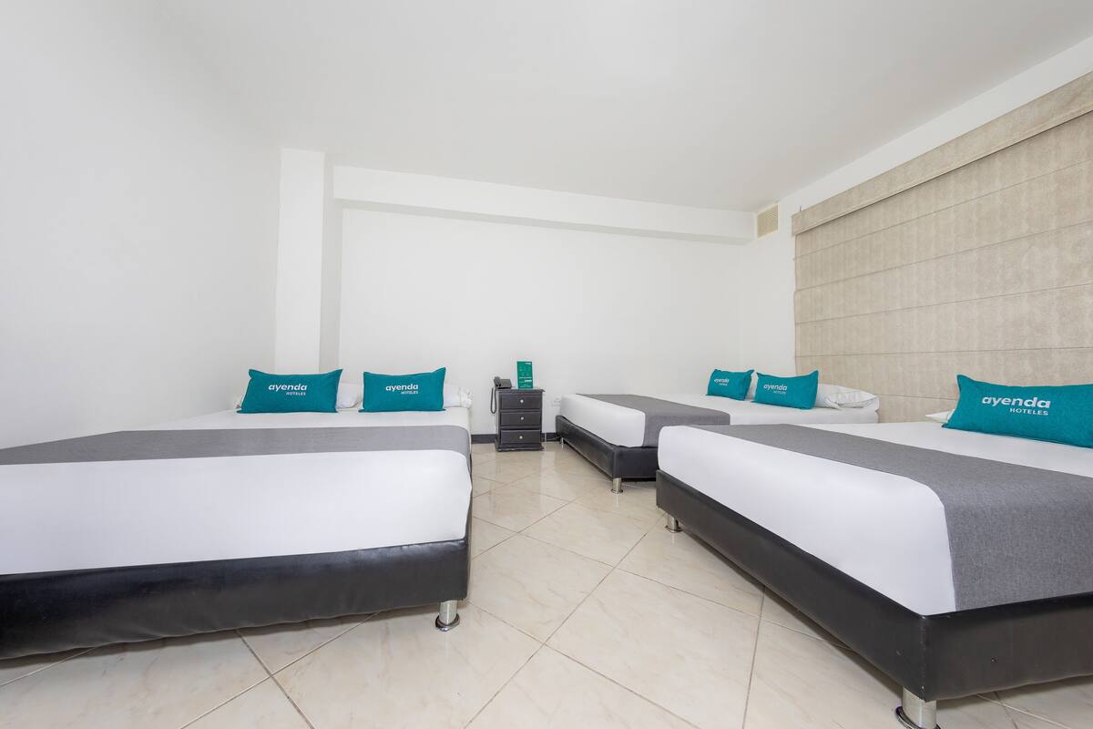A hotel room with three beds, each with teal pillows and a white headboard. There is a small table with a telephone and a green object on it.