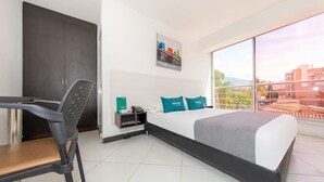 A hotel room with a bed, a desk, a chair, a nightstand, and a large window offering a city view. - Vittapark (Medellín)