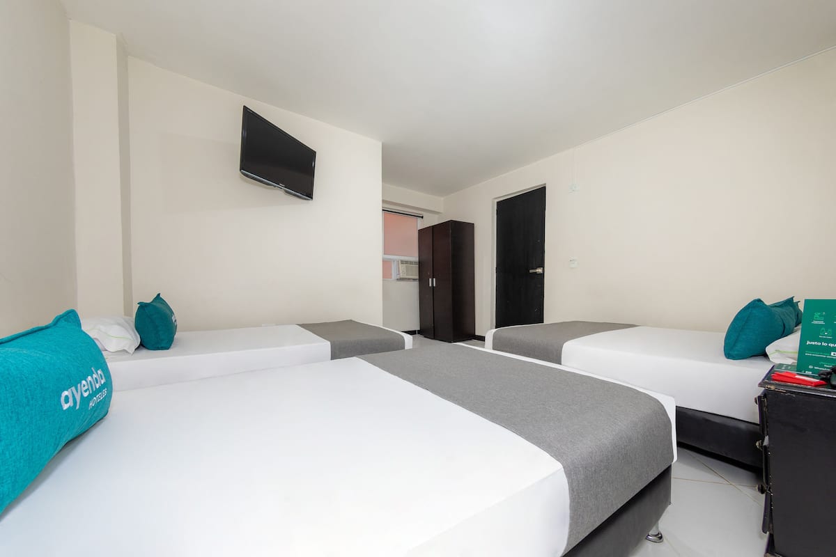 A hotel room with three single beds, a flat-screen TV mounted on the wall, a desk with a computer, and a door leading to a bathroom.