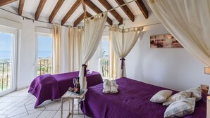 A room with two beds, each with a purple bedspread and white pillows, a small table with a bottle and glasses, and a view of the sea through large windows. - La Torre Cajiz by Ruralidays (Málaga)