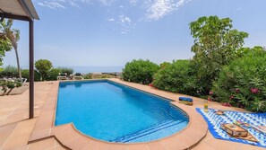 A rectangular swimming pool with a tiled deck, surrounded by lush greenery and a clear view of the sea.