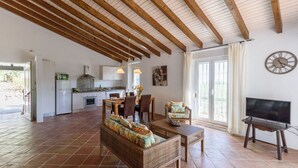 A spacious living area with a dining table, wicker sofa, and a television. A kitchen with a stove and cabinets is visible in the background. - Las Ranas Cajiz by Ruralidays (Málaga)