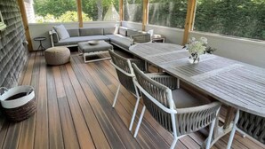 Een overdekt terras met een houten dek, een bank, een eettafel en stoelen.