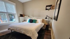 A bedroom with a large bed, a bedside table, a nightstand, a lamp, a window with blinds, and a wall-mounted picture frame. - Long Lake Oasis: To recharge or Work with a view (Lacey)