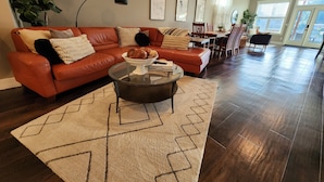 A spacious living room with a red leather sofa, a glass coffee table, and a patterned rug. There are framed pictures on the wall, a dining area with wooden chairs, and a large window providing natural light. - Long Lake Oasis: To recharge or Work with a view (Lacey)