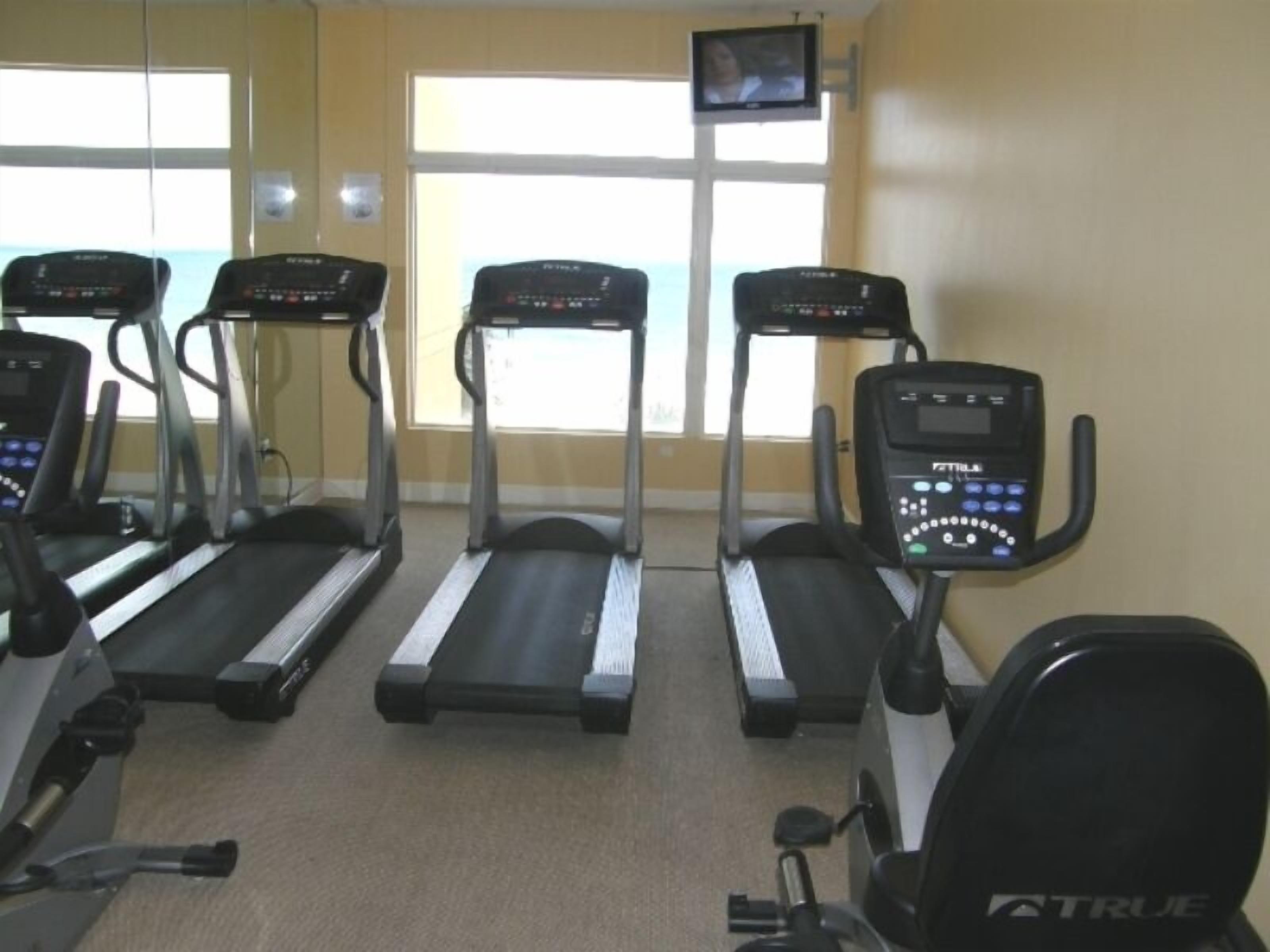A gym with treadmills, a TV mounted on the wall, and large windows.