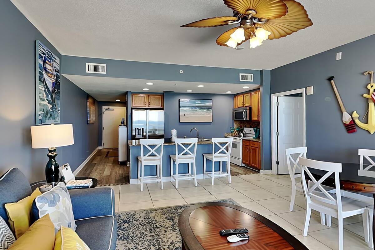 A modern living room with a sofa, a coffee table, and a TV. There is a kitchen area with a bar and white chairs.