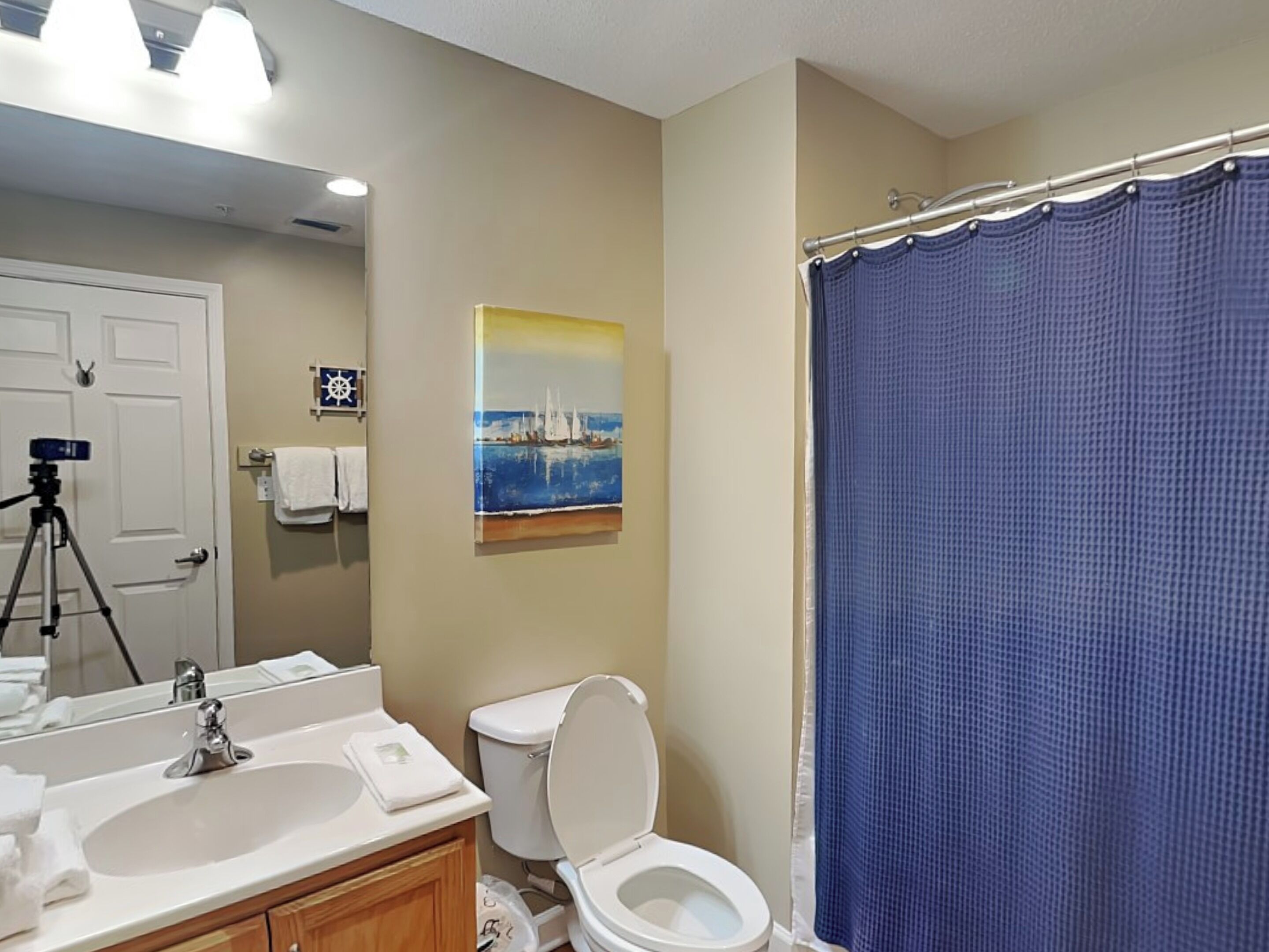 A bathroom with a white sink, a toilet, a blue shower curtain, and a painting of a sailboat on the wall.